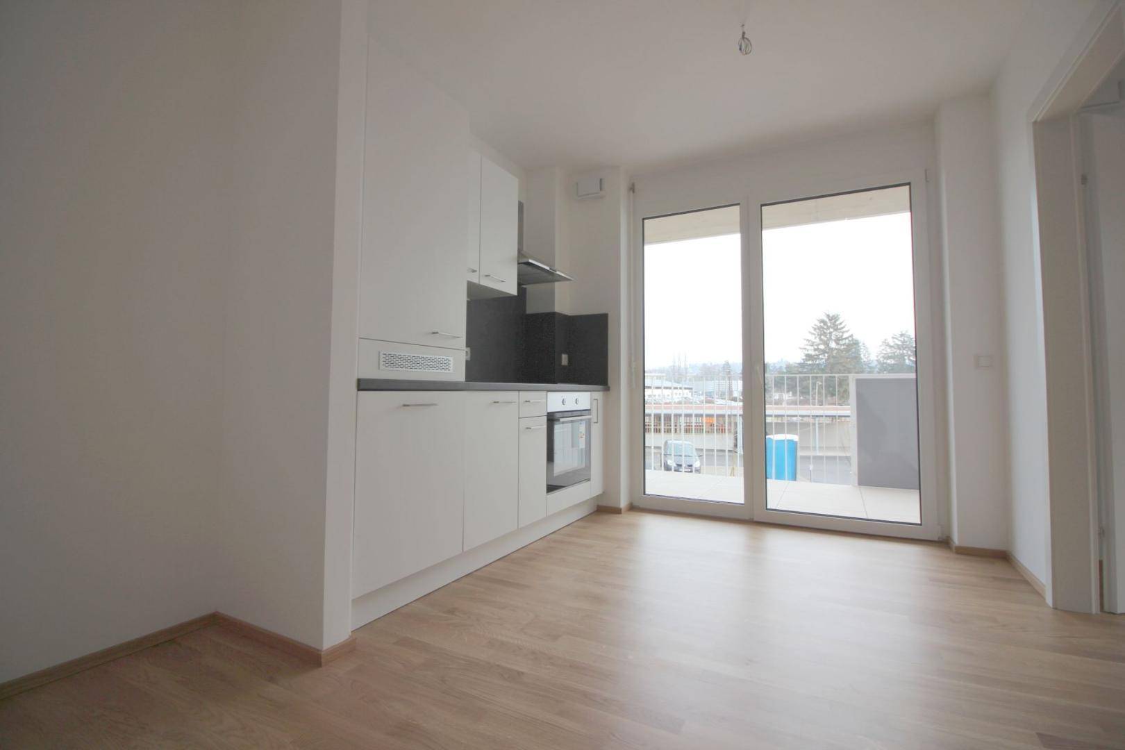 Heller offener Wohnbereich mit integrierter Küchenzeile, Parkettboden und Zugang zum Balkon.