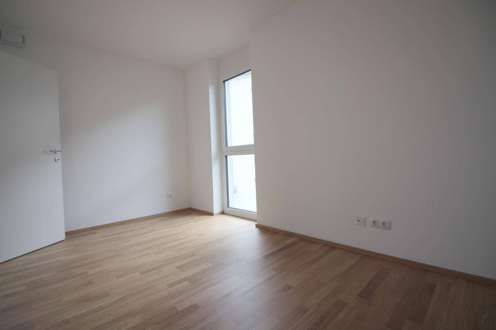 Helles Zimmer mit Parkettboden und einem hohen Fenster, bereit zur individuellen Gestaltung.
