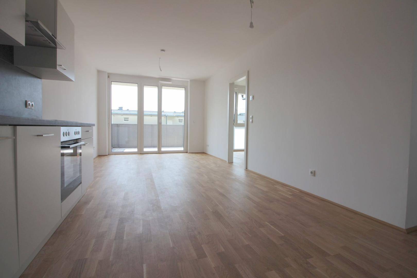 Heller offener Wohnbereich mit Parkettboden, Küchenzeile und Zugang zum Balkon.