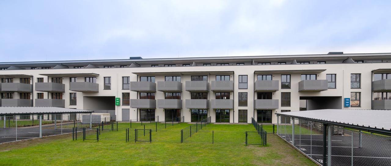 Lange Gebäudefassade mit Balkonen, Grünflächen und überdachten Parkplätzen.