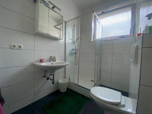 Funktionales Badezimmer mit weißen Fliesen, Duschkabine, Waschbecken und einem Fenster für Belüftung.