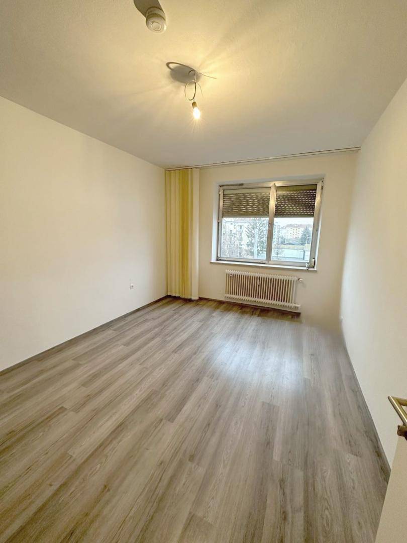 Helles Schlafzimmer mit Laminatboden, weiß gestrichenen Wänden und einem Fenster mit Heizkörper.