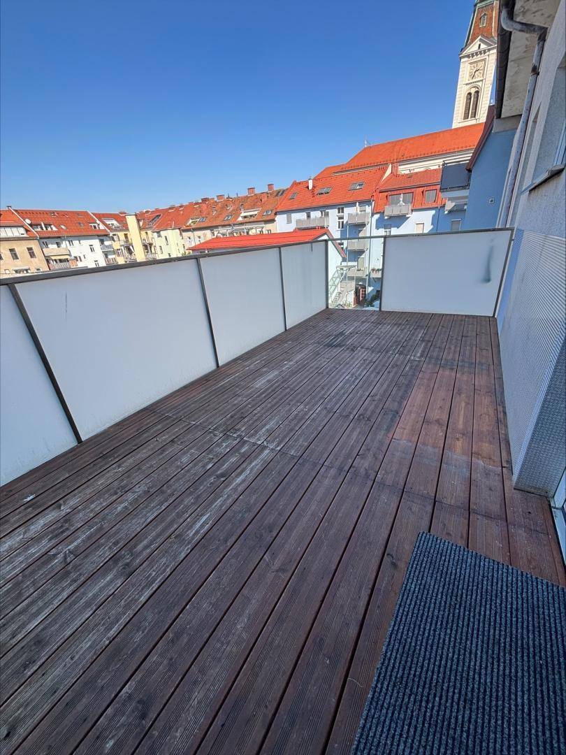 Geräumiger Balkon mit Holzboden und Blick auf die Dächer der Stadt und einen Kirchturm.