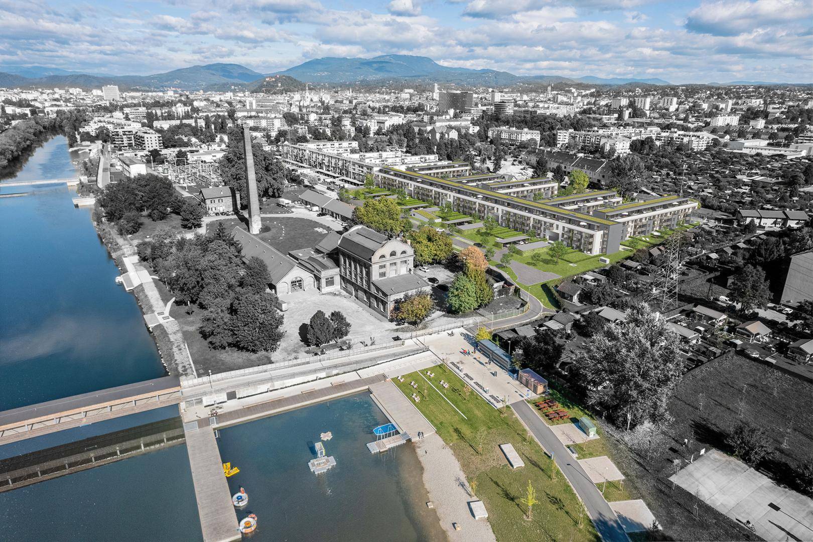 Luftaufnahme einer Stadt mit Fluss, Brücke und neuer Wohnbebauung vor Bergkulisse.