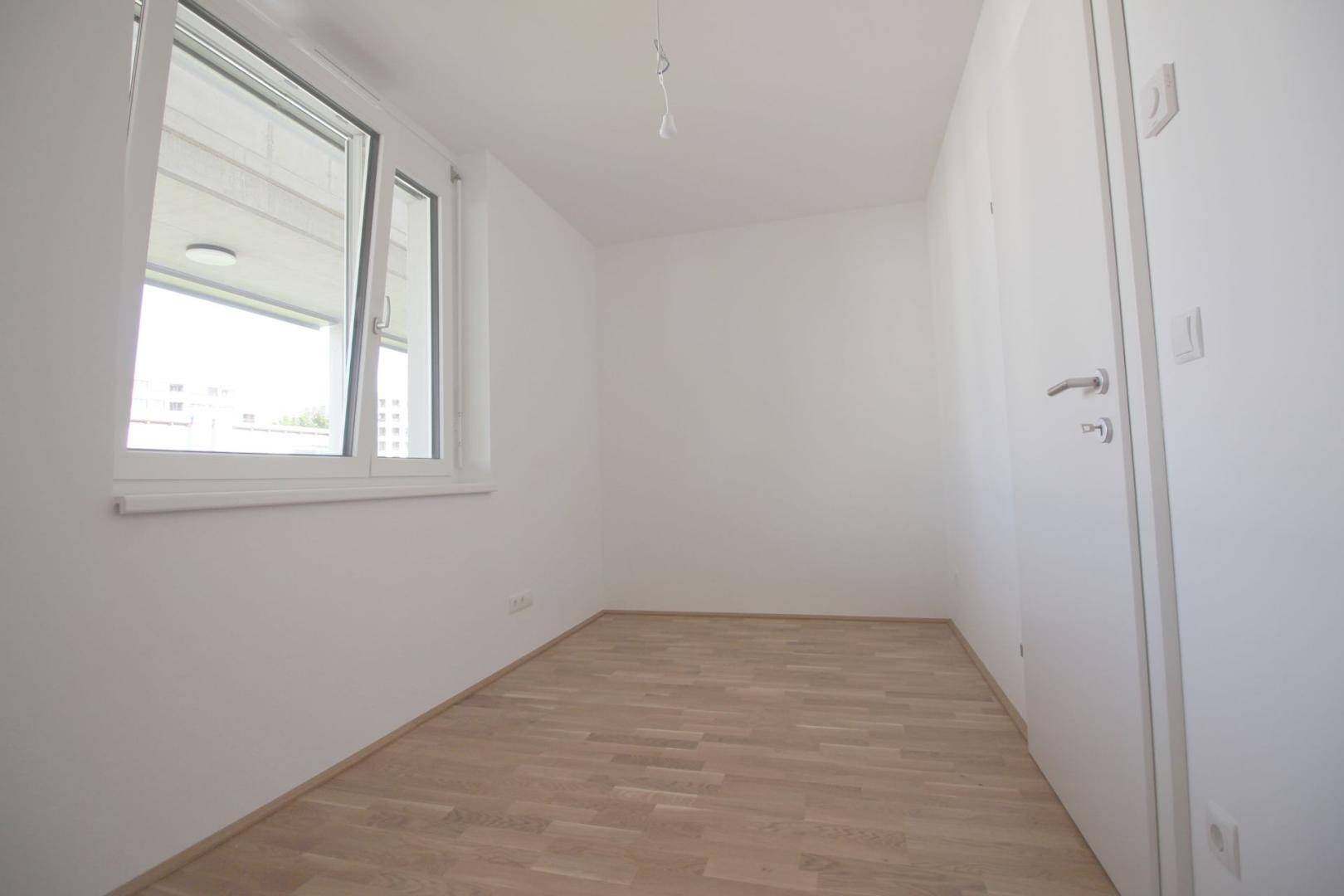 Kleiner, heller Raum mit Fenster, hellem Parkettboden und weißer Tür.