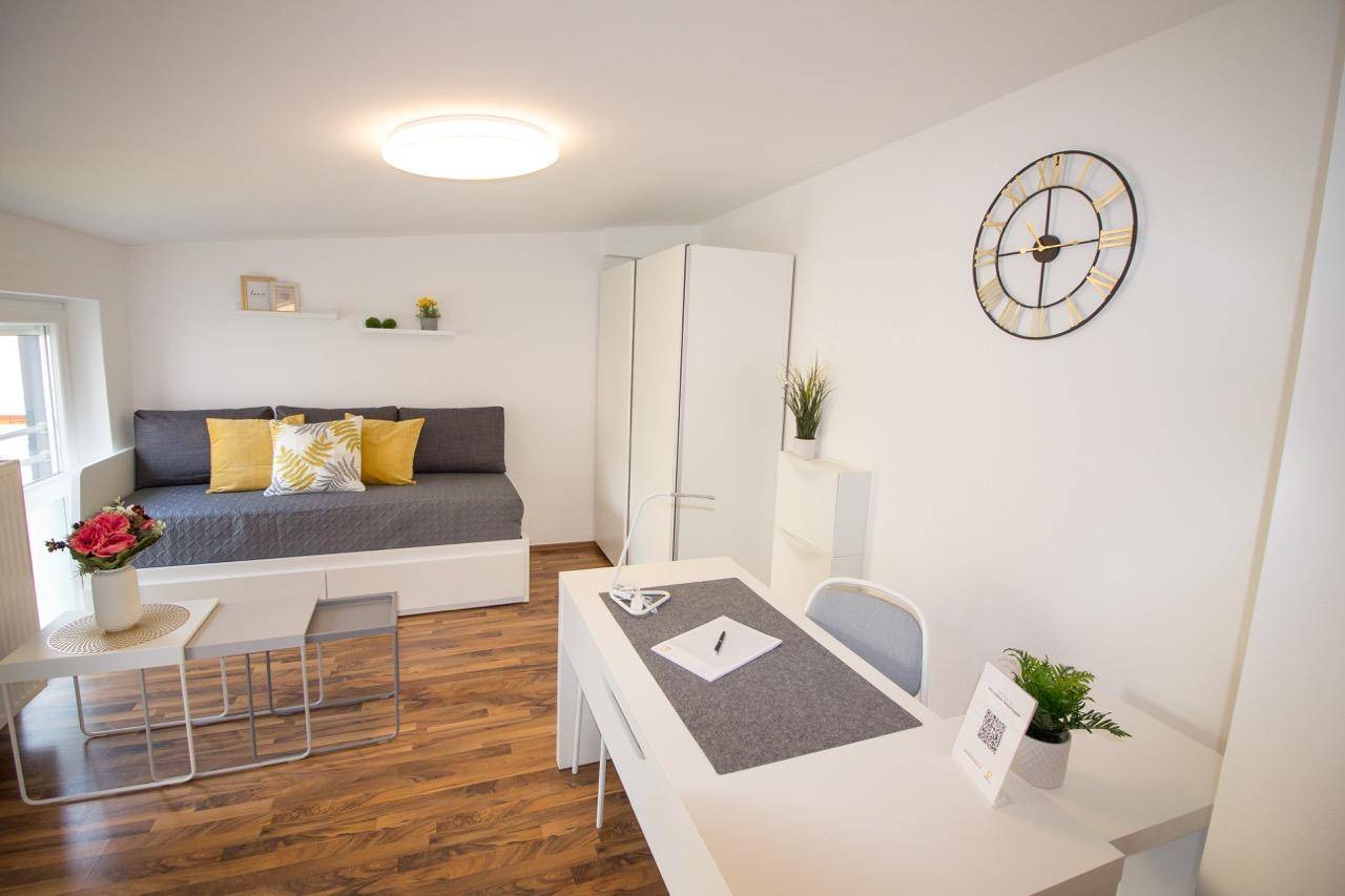 Gemütlicher Wohnbereich mit Schlafsofa, Holzboden und einer modernen Wanduhr als Dekoration.