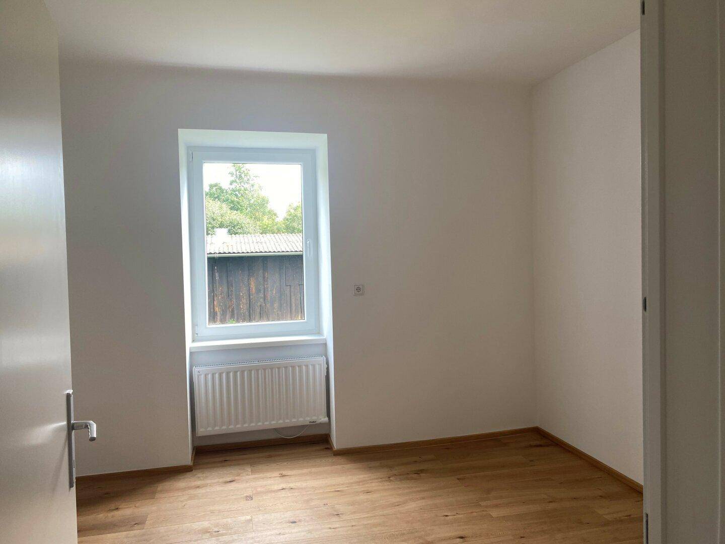 Leeres Zimmer mit Holzboden und Fenster, das viel Tageslicht hereinlässt und einen grünen Ausblick bietet.