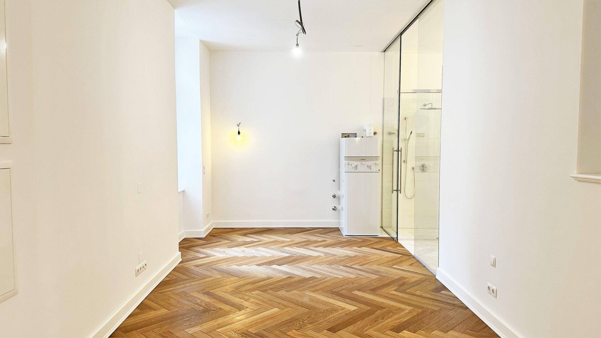 Raum mit Fischgrätparkett und einer modernen Glasduschkabine, daneben ein Kühlschrank.