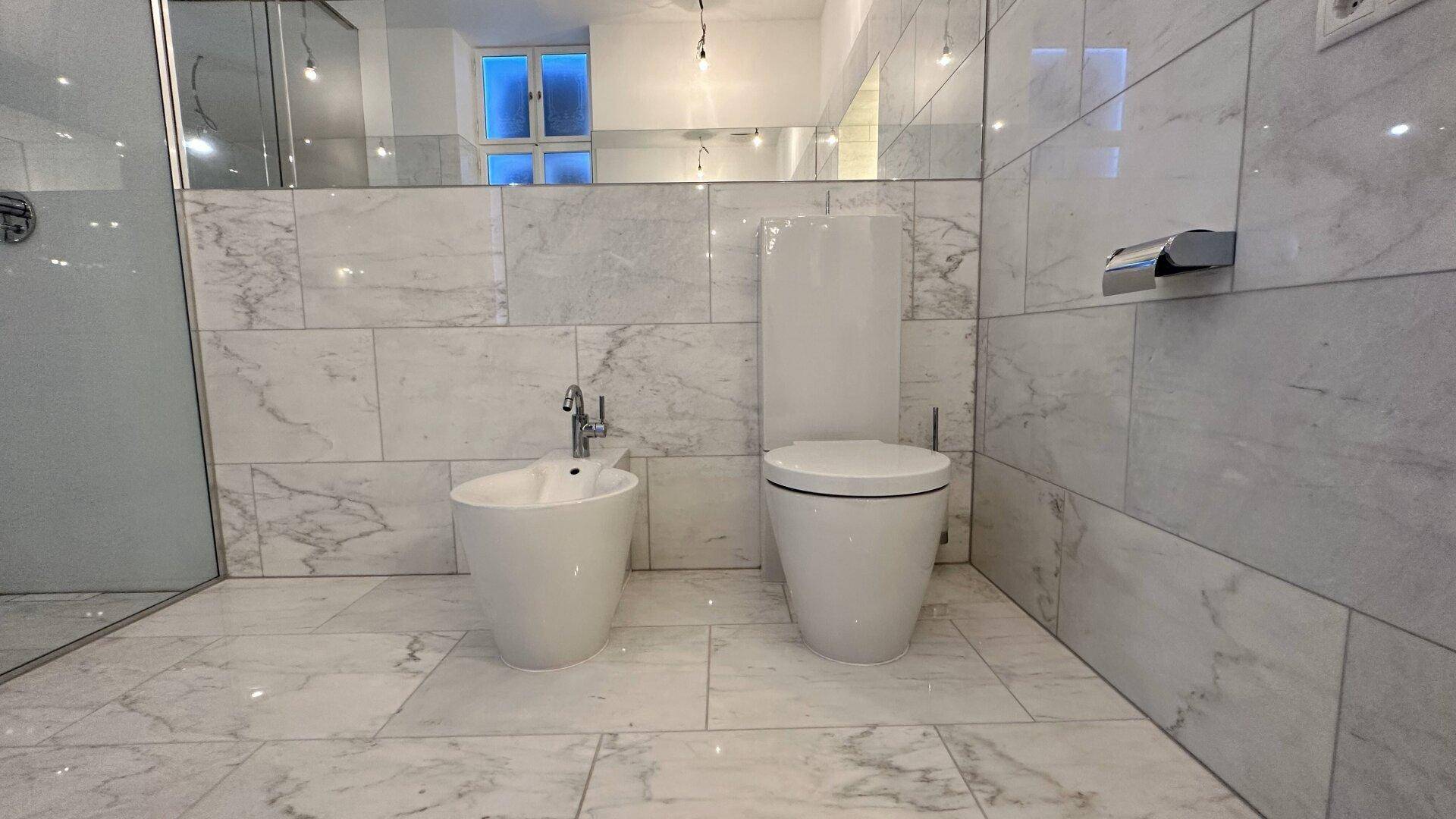 Badezimmer mit Marmorfliesen, wandhängendem WC und Bidet, sowie einem großen Spiegel.