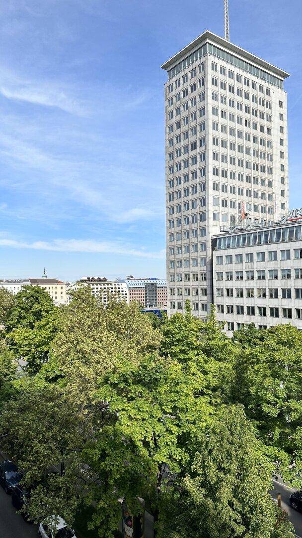Weitläufiger Blick auf die Stadt mit einem modernen Hochhaus und vielen grünen Bäumen.