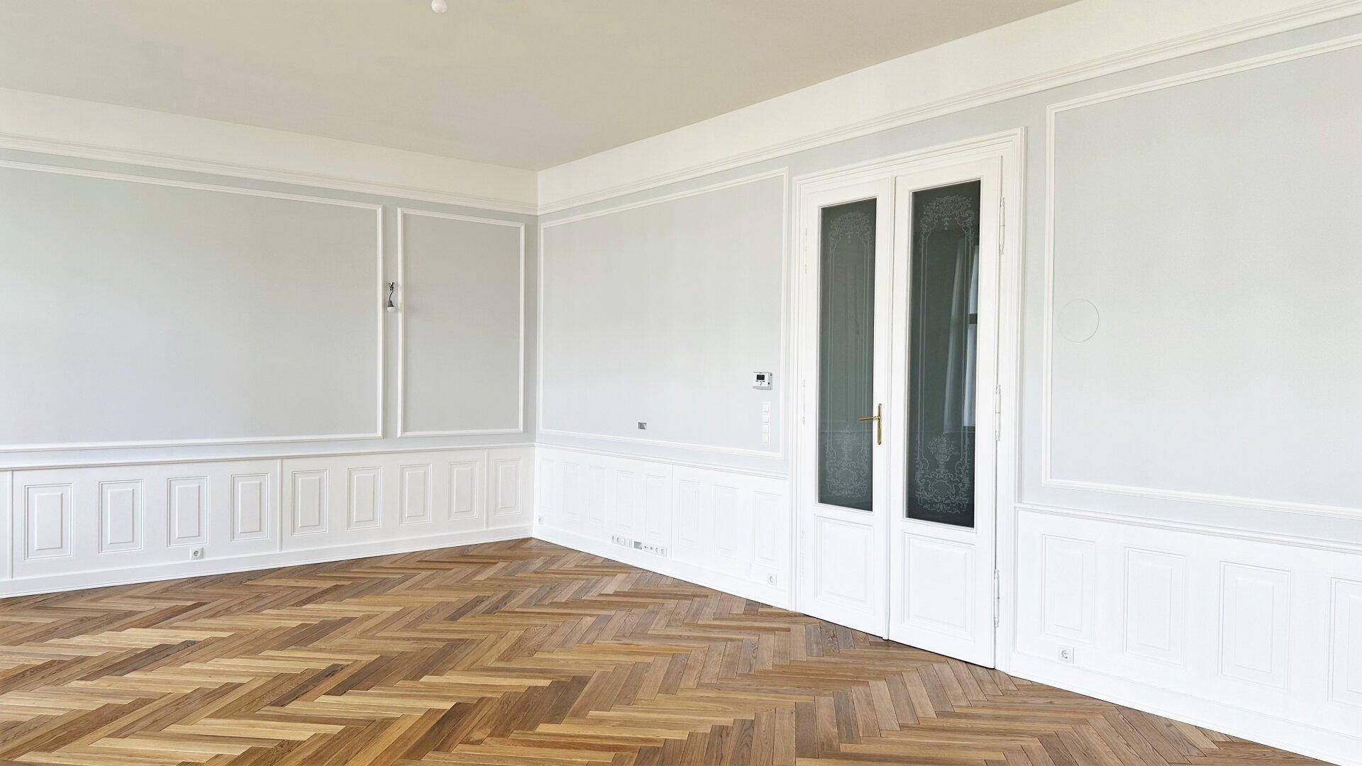 Elegantes Zimmer mit Fischgrätparkett, hohen Decken und einer Doppeltür mit Glasausschnitten.