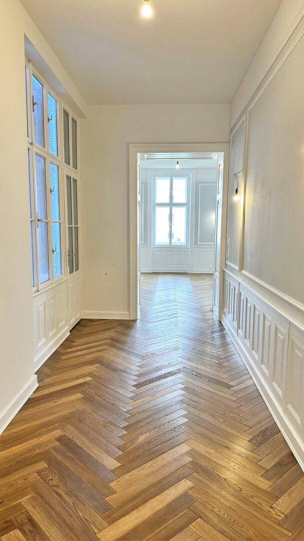 Heller Flur mit Fischgrätparkett und Blick in ein weiteres Zimmer mit hohen Fenstern.