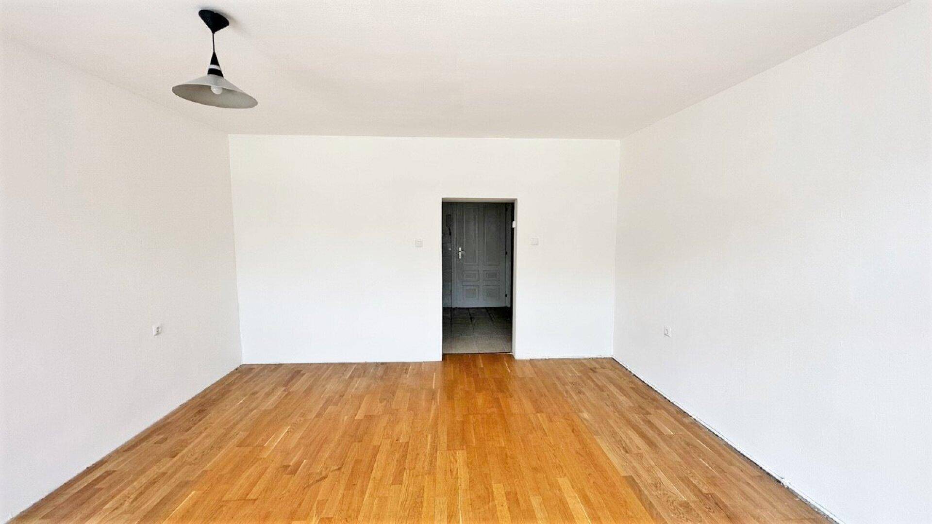 Leeres Wohnzimmer mit Parkettboden und einem offenen Durchgang zu einem weiteren Raum.