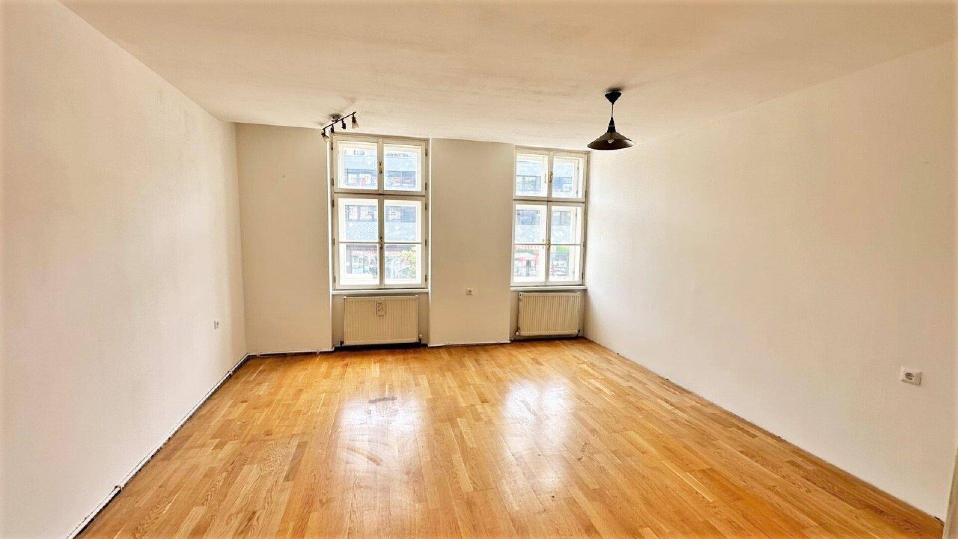 Geräumiges, unmöbliertes Wohnzimmer mit hellem Parkettboden und zwei Fenstern.