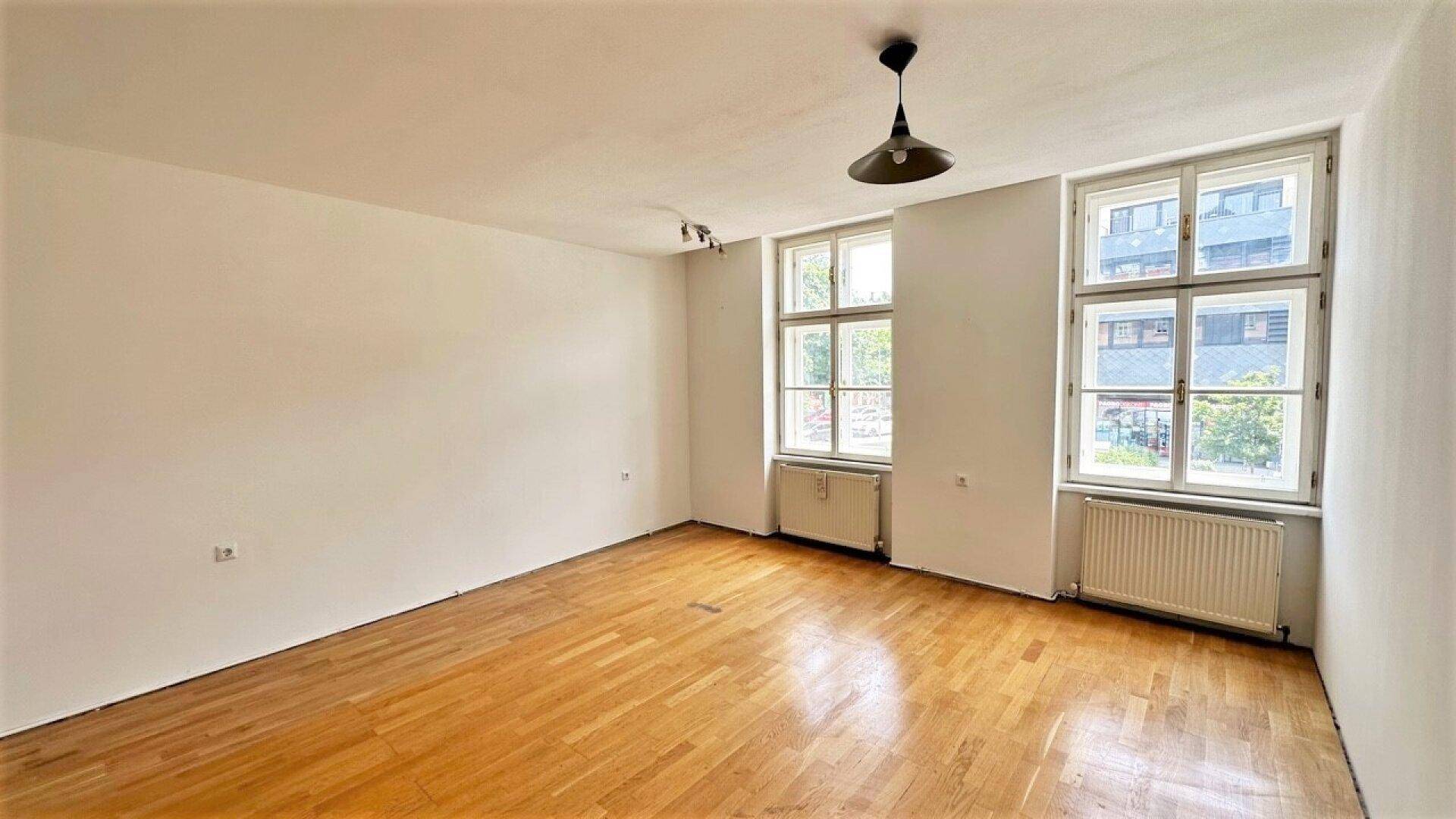 Helles Wohnzimmer mit Parkettboden und zwei Fenstern, die Tageslicht hereinlassen.