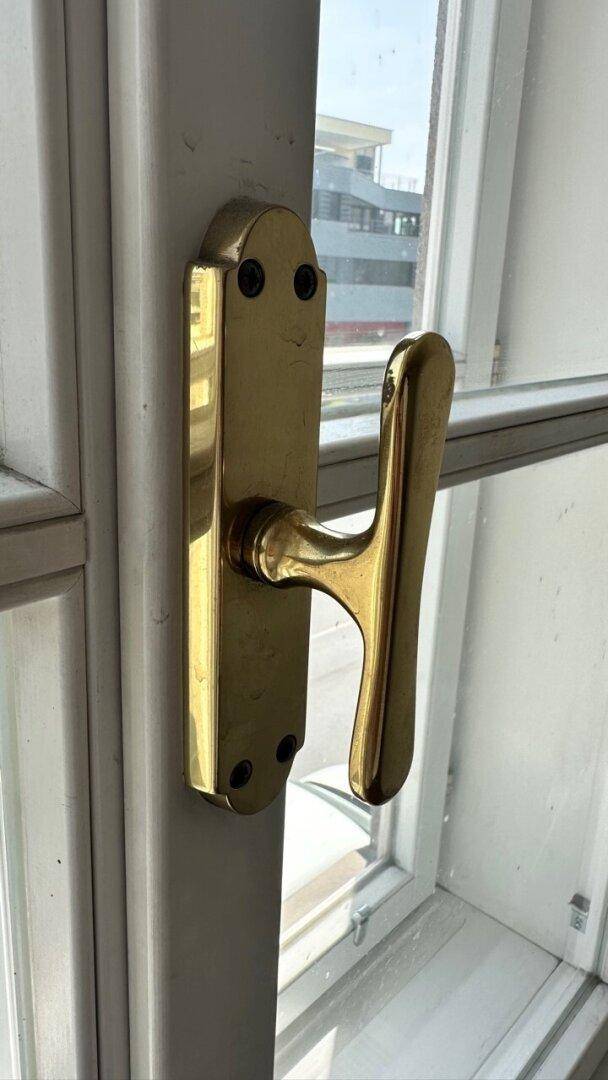 Nahaufnahme eines goldenen Fenstergriffs an einem weißen Fensterrahmen.