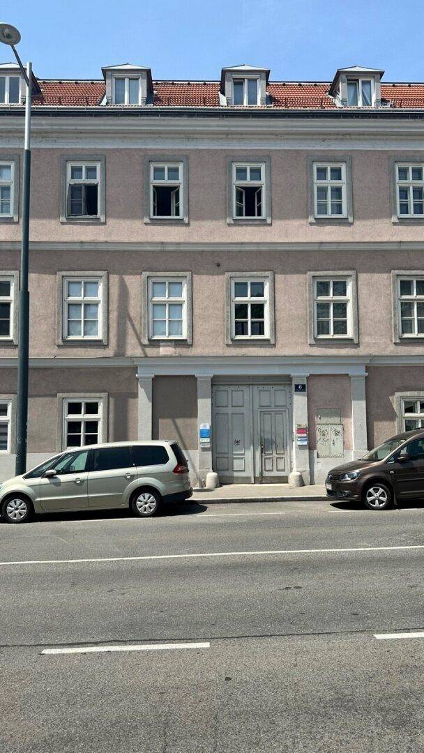 Fassade eines Altbaus mit vielen Fenstern und parkenden Autos entlang der Straße.