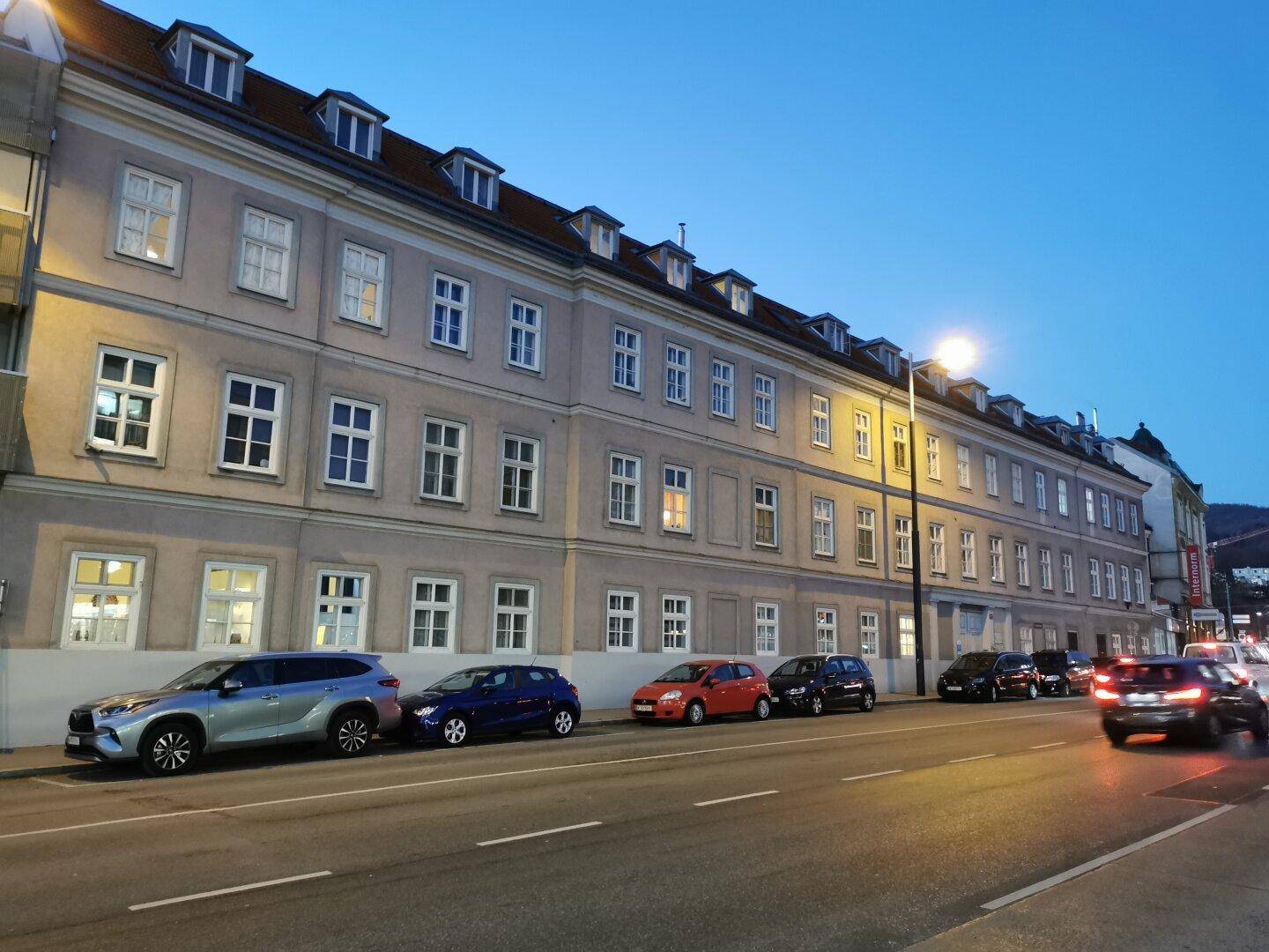 Historisches Gebäude bei Dämmerung mit beleuchteten Fenstern und parkenden Autos.
