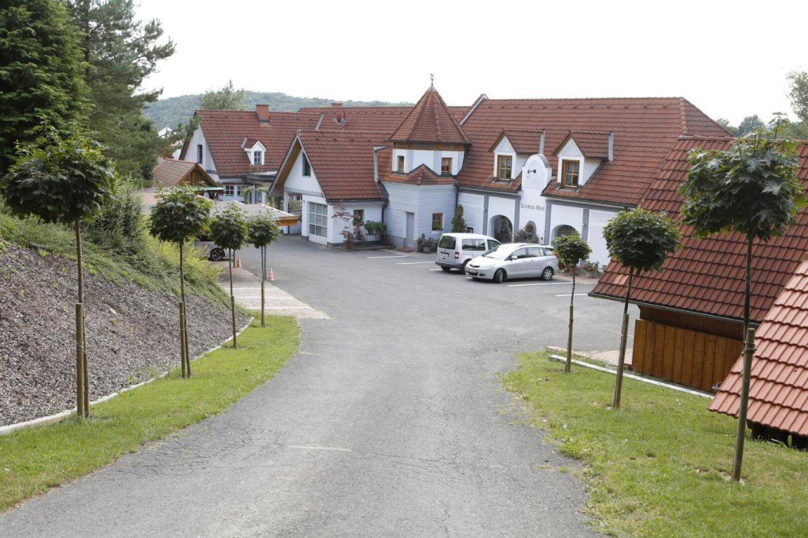 Ansicht des Gebäudes mit Parkplatz und Zufahrt, umgeben von Bäumen und gepflegter Landschaft.