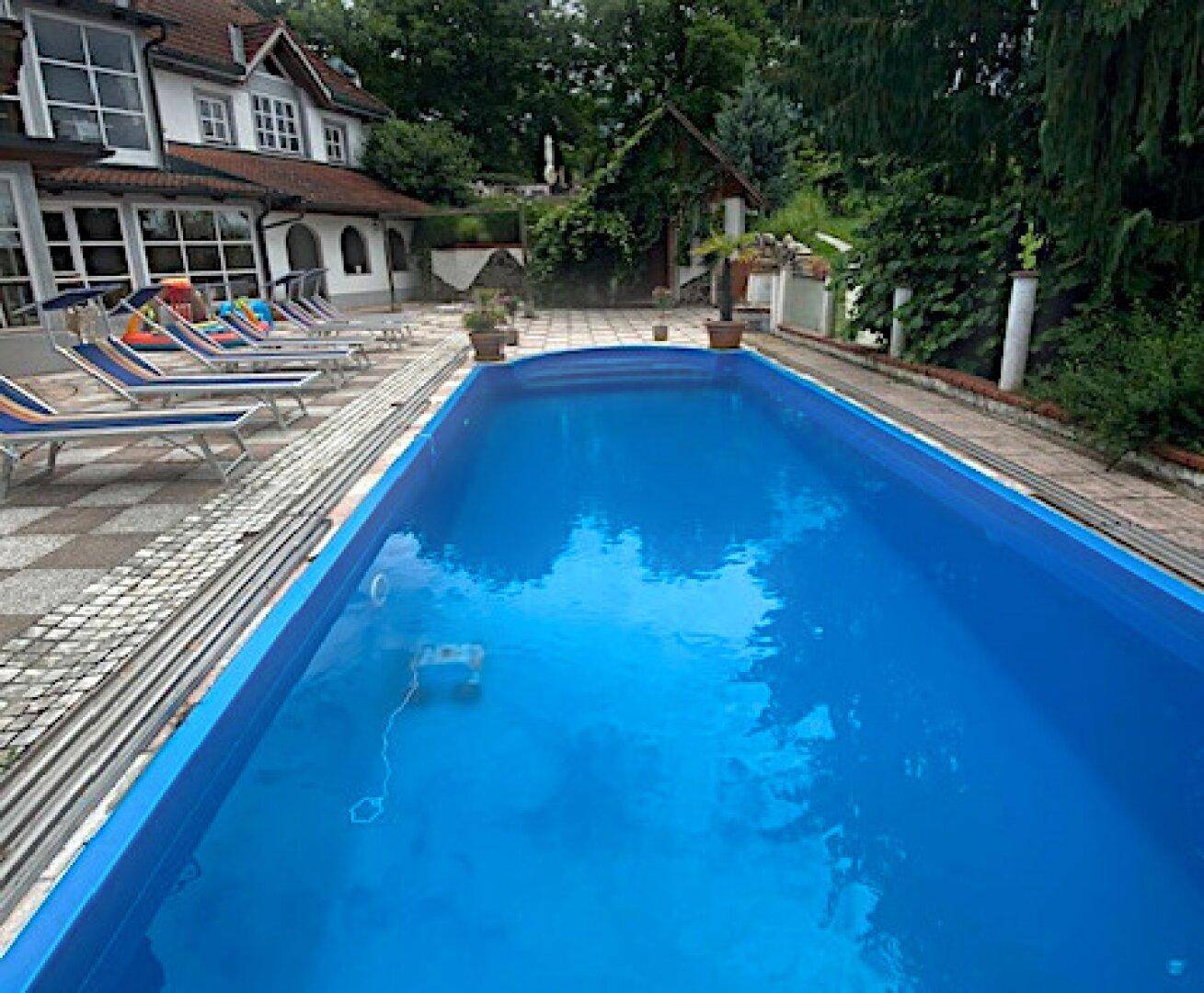Außenpoolbereich mit Liegestühlen und gepflasterter Terrasse, umgeben von Grün.
