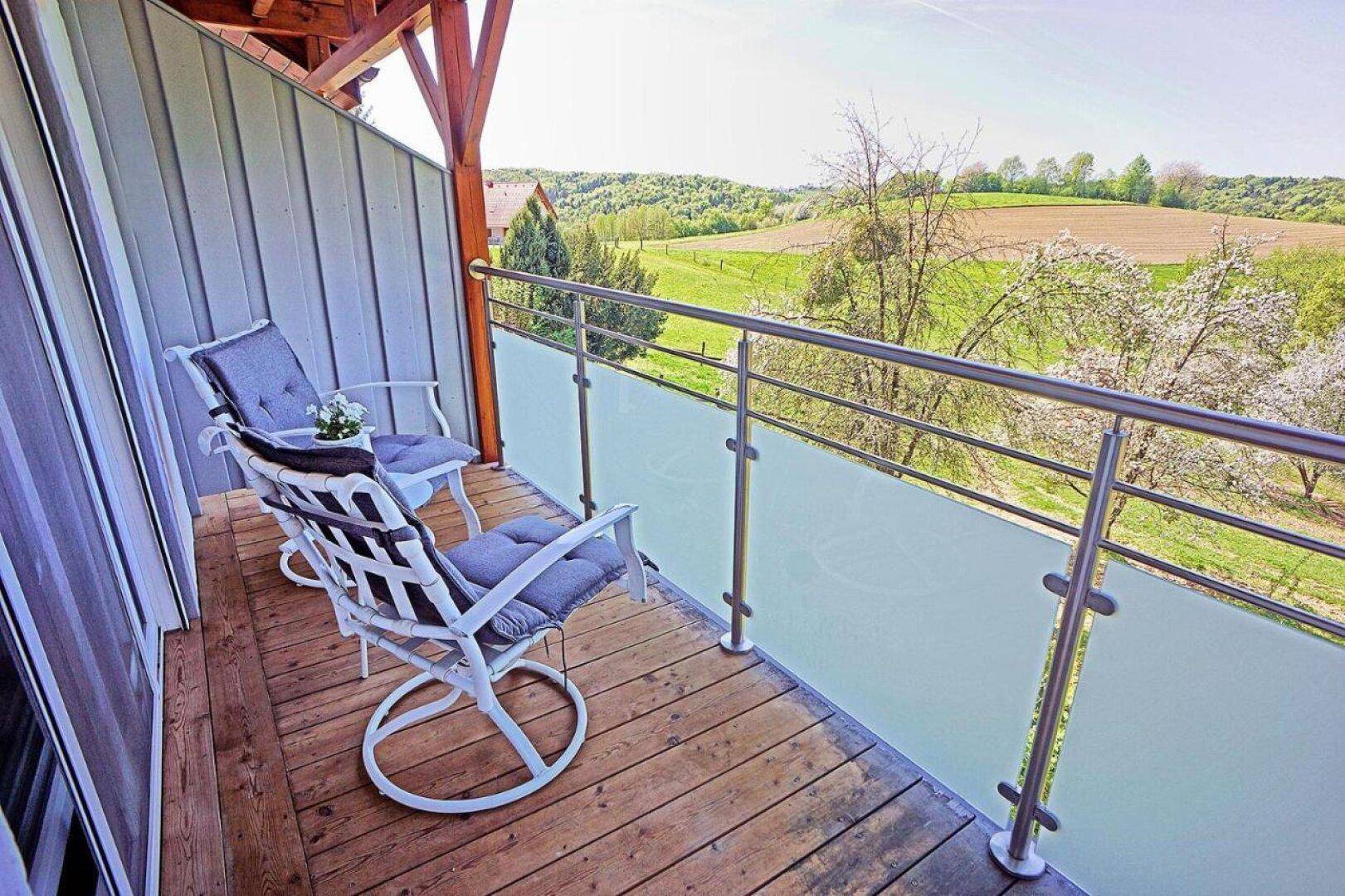 Balkon mit Holzboden und Glasgeländer, möbliert mit Stühlen und weitem Blick über die Landschaft.