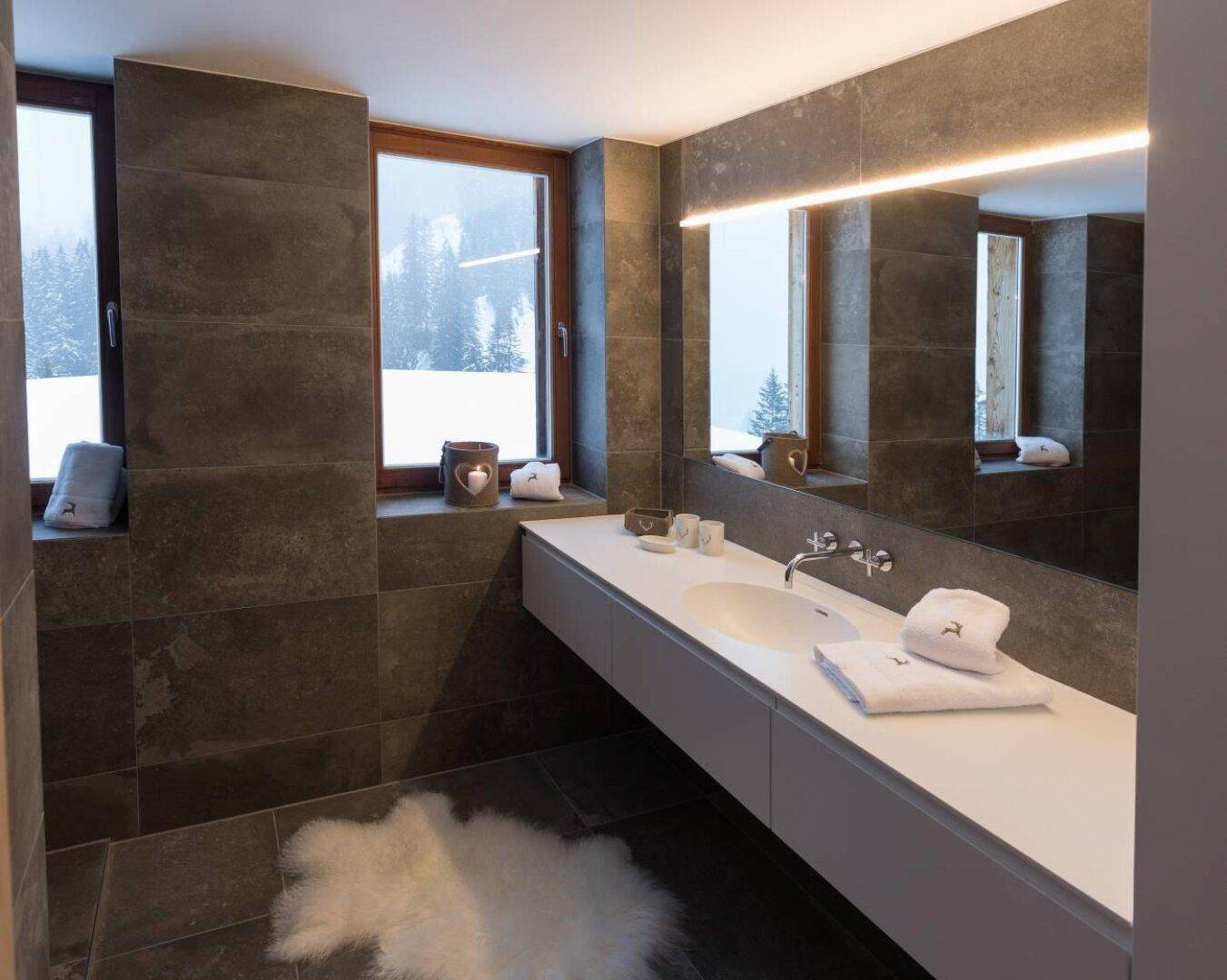 Modernes Badezimmer mit dunklen Fliesen, Doppelwaschbecken und Blick auf die verschneite Landschaft.