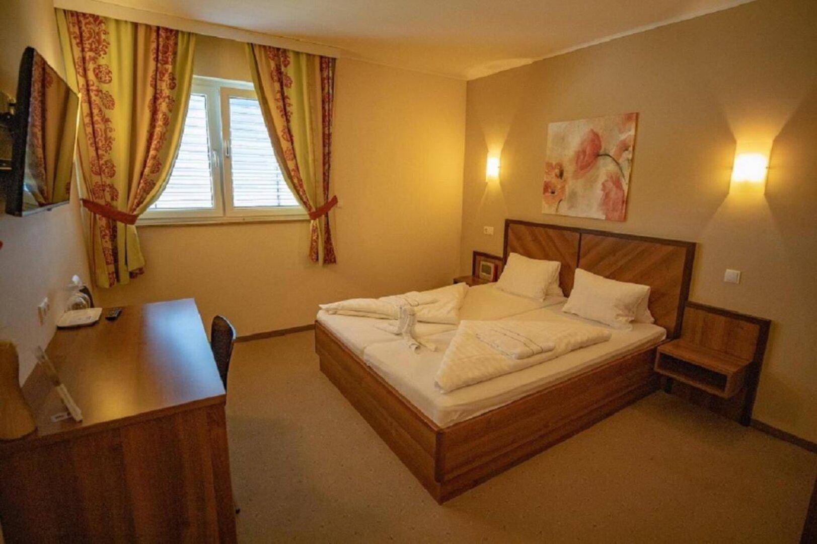 Gemütliches Schlafzimmer mit Doppelbett, Holzmöbeln und einem Schreibtisch neben dem Fenster mit Vorhängen.