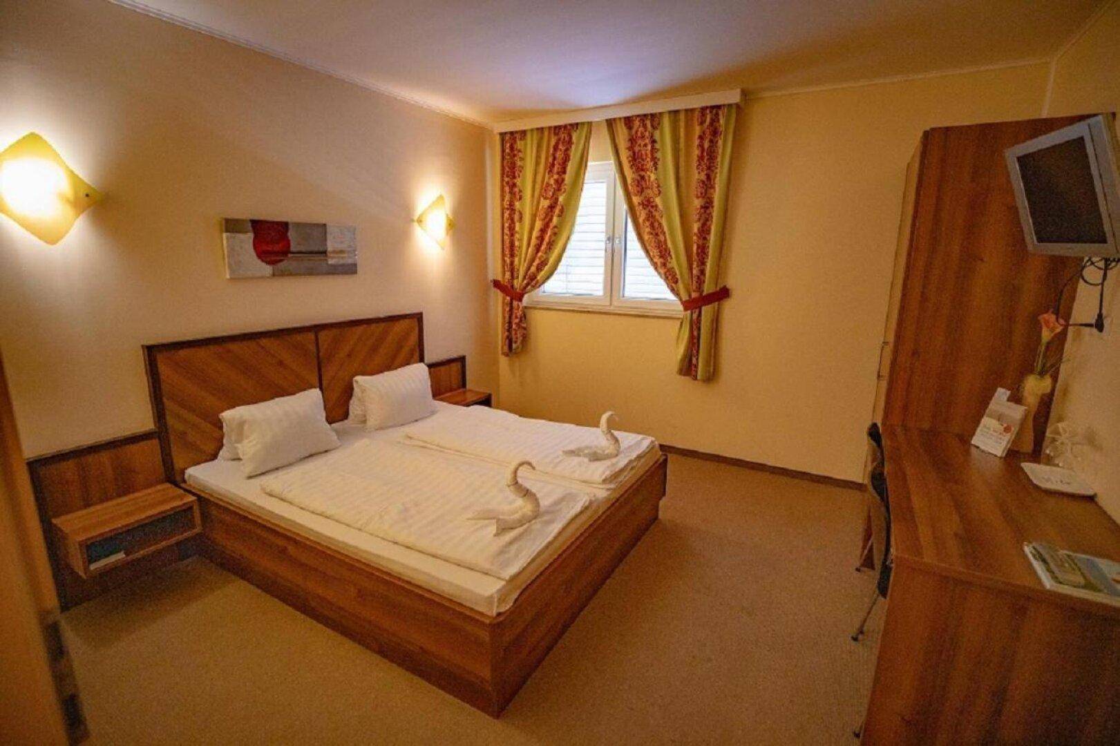 Helles Schlafzimmer mit Doppelbett, Holzschrank und einem Schreibtisch neben dem Fenster mit bunten Vorhängen.
