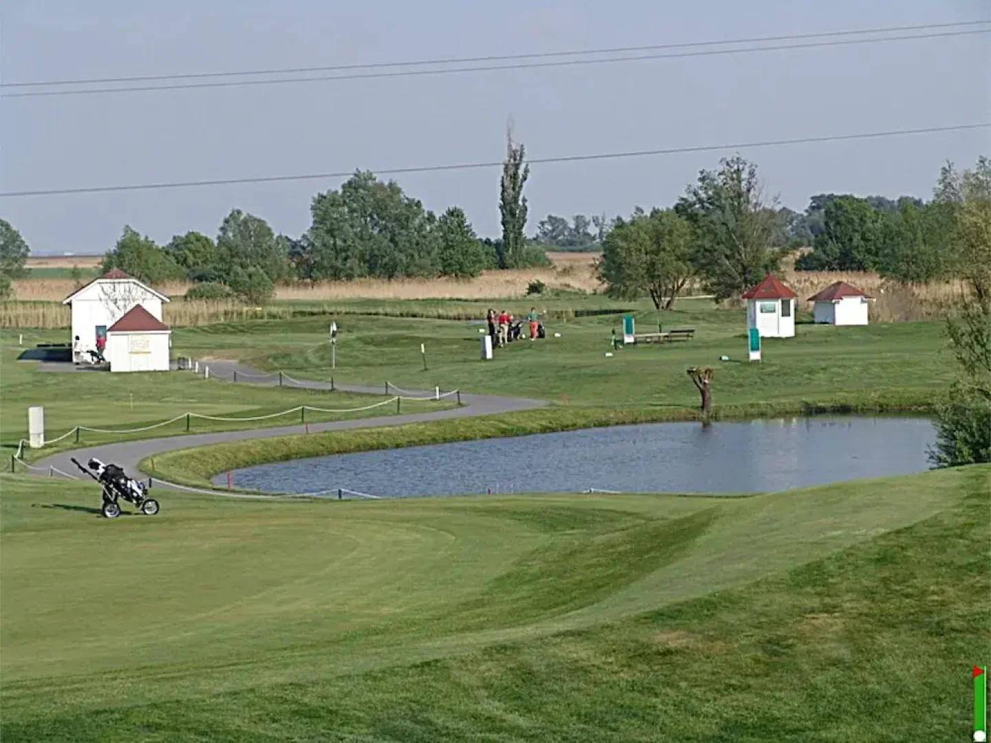 Weitläufiger Golfplatz mit einem ruhigen Teich, grünen Fairways und kleinen Unterständen unter klarem Himmel.