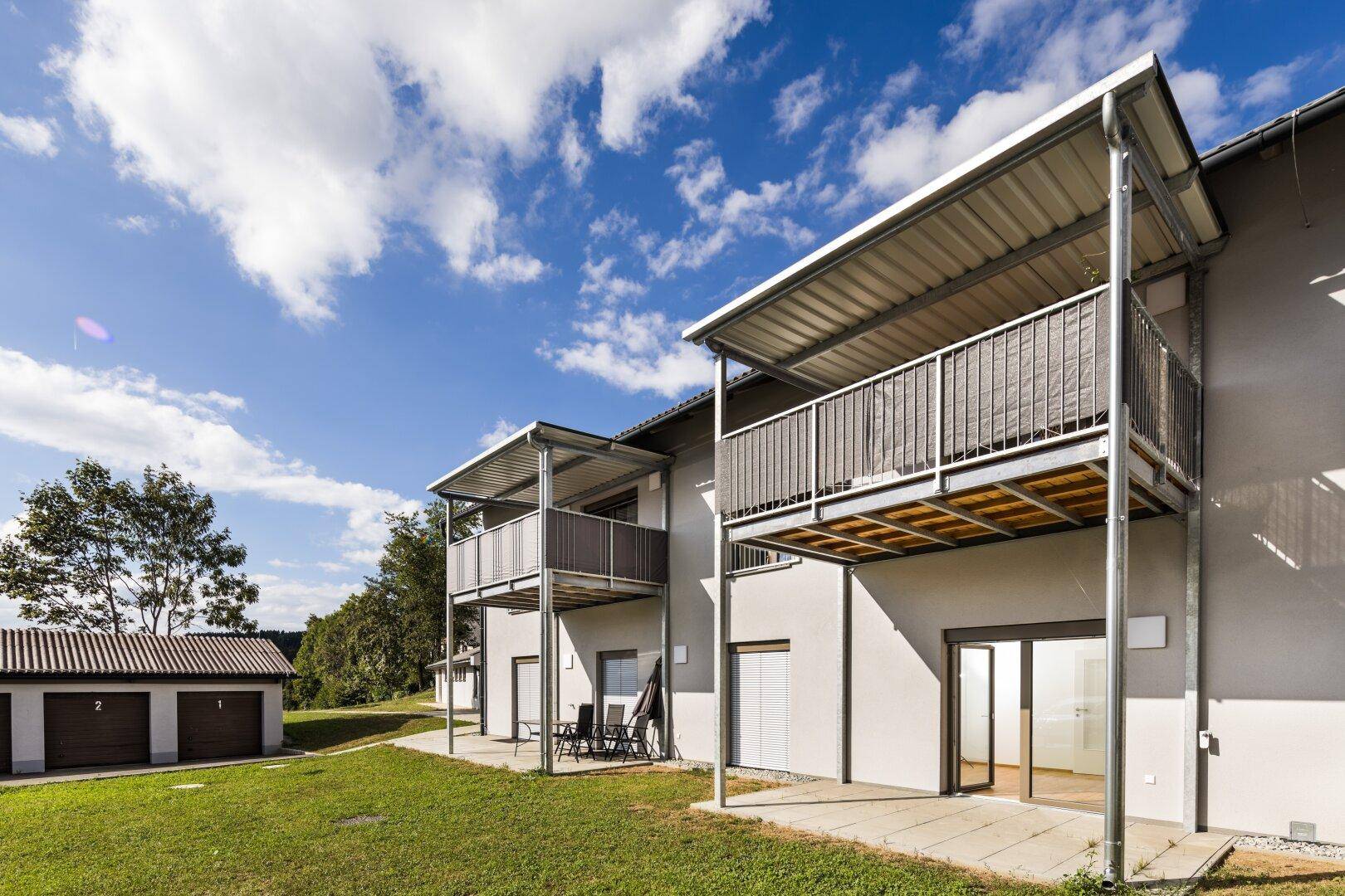 Ansicht eines modernen Mehrfamilienhauses mit Balkonen und angrenzender Garage in ländlicher Umgebung.