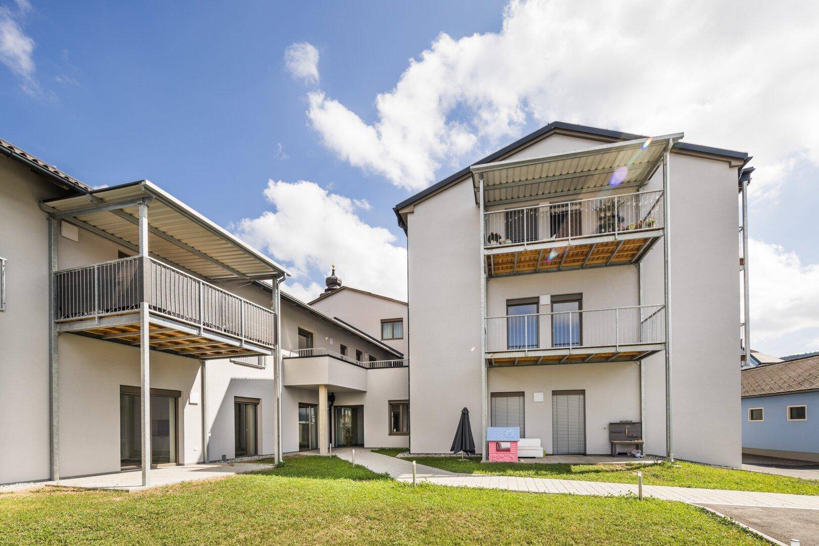 Modernes Mehrfamilienhaus mit mehreren Balkonen und gepflegten Grünflächen unter blauem Himmel.