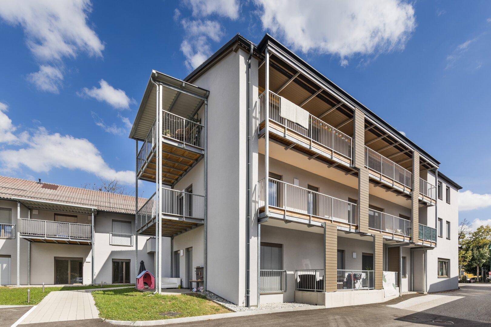 Seitenansicht eines modernen Mehrfamilienhauses mit mehreren Balkonen und angrenzendem Parkplatz.