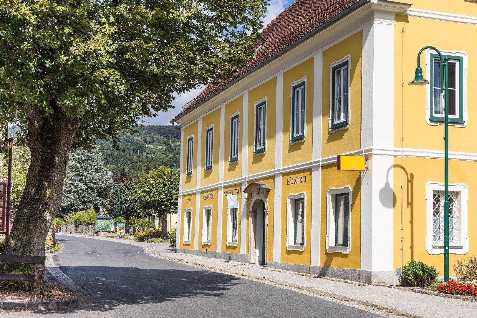 Gelbes historisches Gebäude mit grünen Fensterrahmen an einer Dorfstraße.