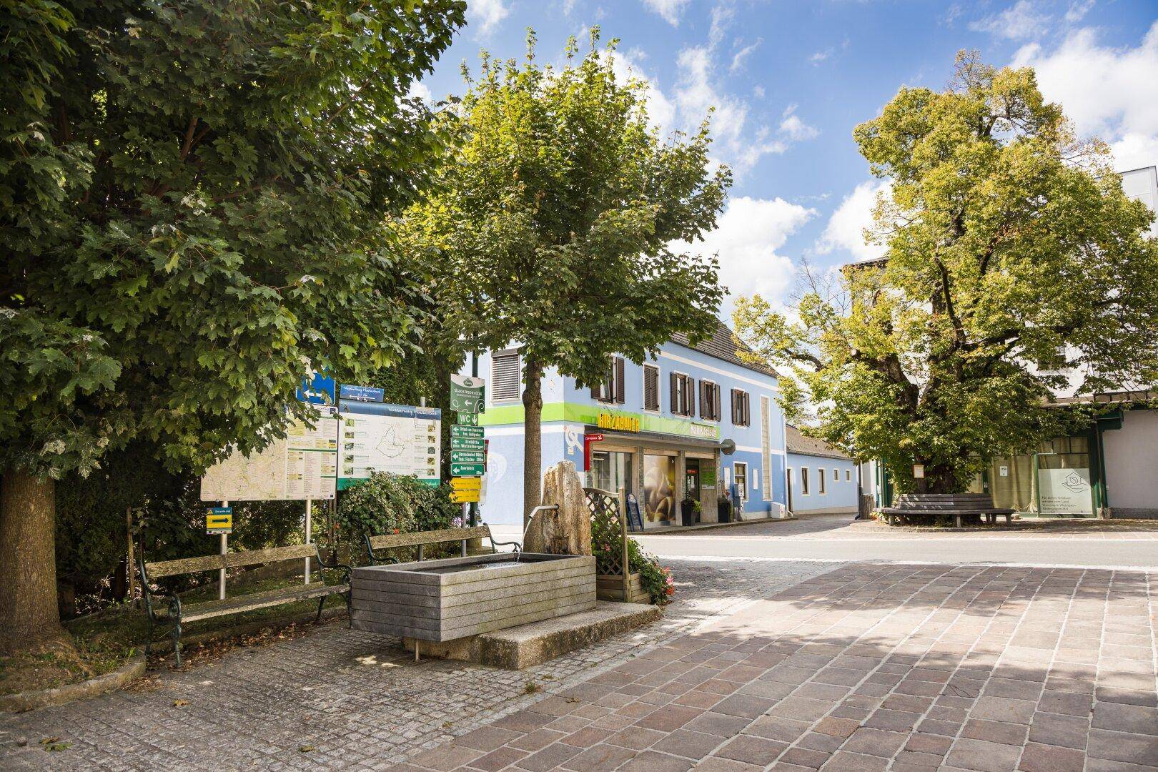 Dorfplatz mit Brunnen, Bäumen und traditionellen Gebäuden unter blauem Himmel.
