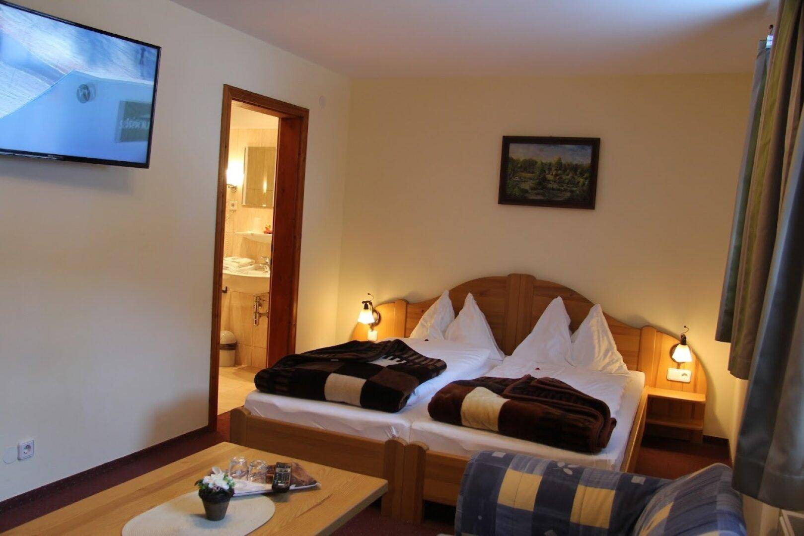 Gemütliches Schlafzimmer mit zwei Einzelbetten, einem Fernseher an der Wand und Blick auf ein angrenzendes Badezimmer.