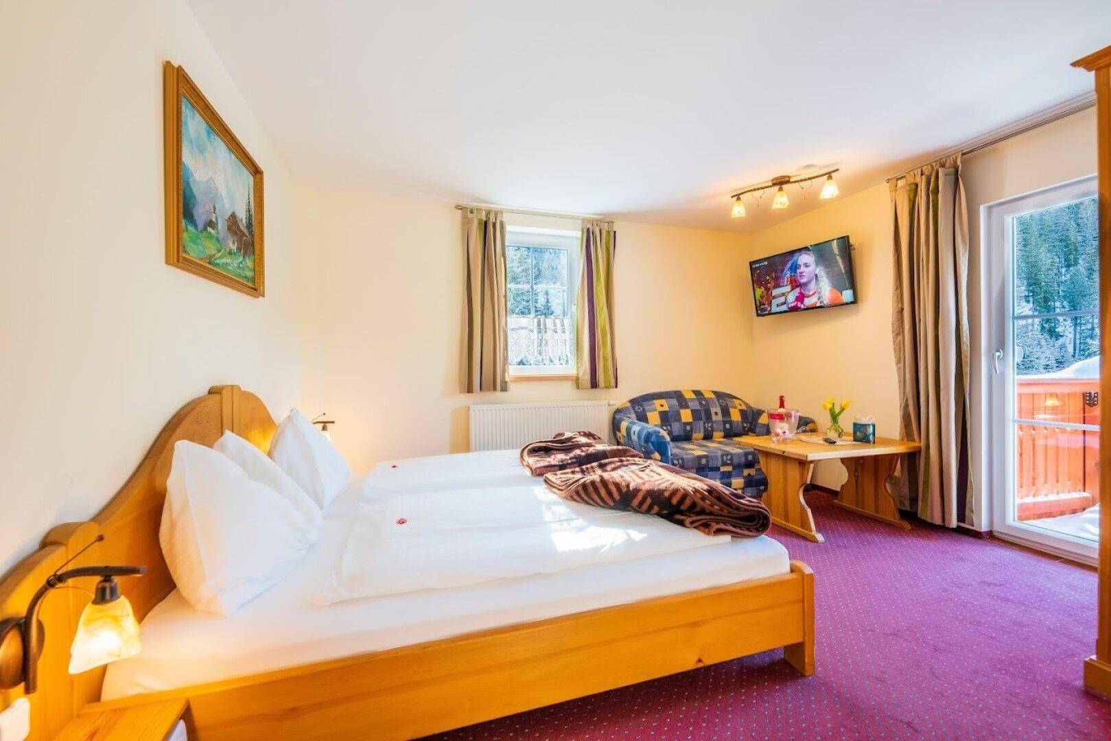 Helles Schlafzimmer mit zwei Einzelbetten, einem Sofa, Fernseher und Zugang zu einem Balkon mit Winterlandschaft.