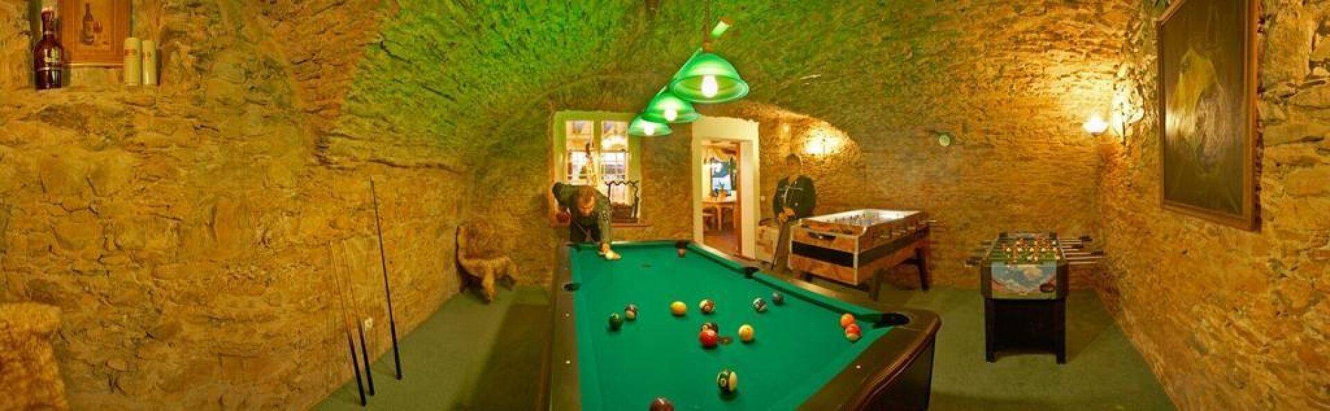 Rustikaler Spielraum mit Billardtisch, Tischfußball und unverputzten Steinwänden, beleuchtet von grünen Lampen.