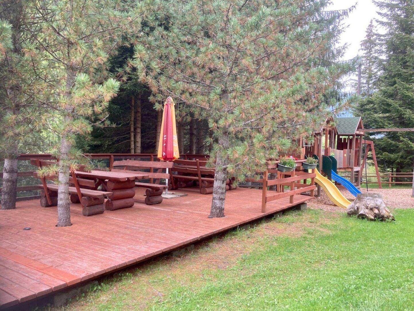 Große Holzterrasse mit rustikalen Sitzgelegenheiten und einem Kinderspielplatz im Hintergrund, umgeben von Bäumen.