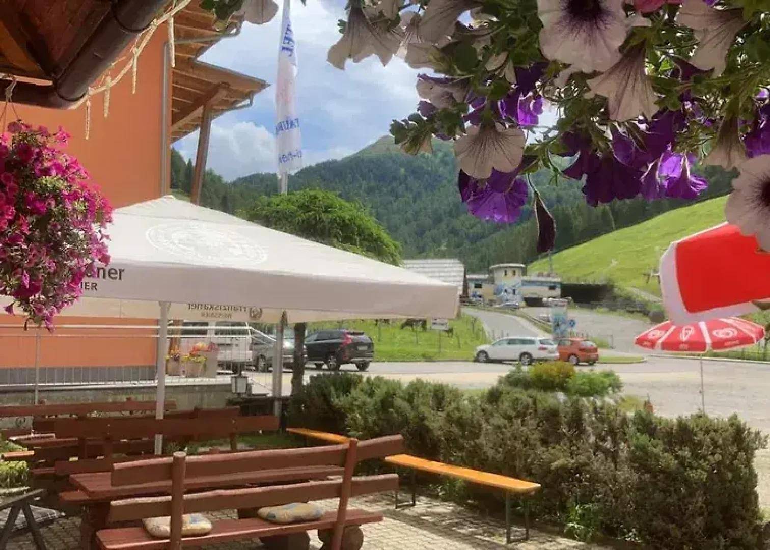 Gemütlicher Außenbereich mit Holzbänken, Sonnenschirmen und blühenden Blumen vor einer Bergkulisse.