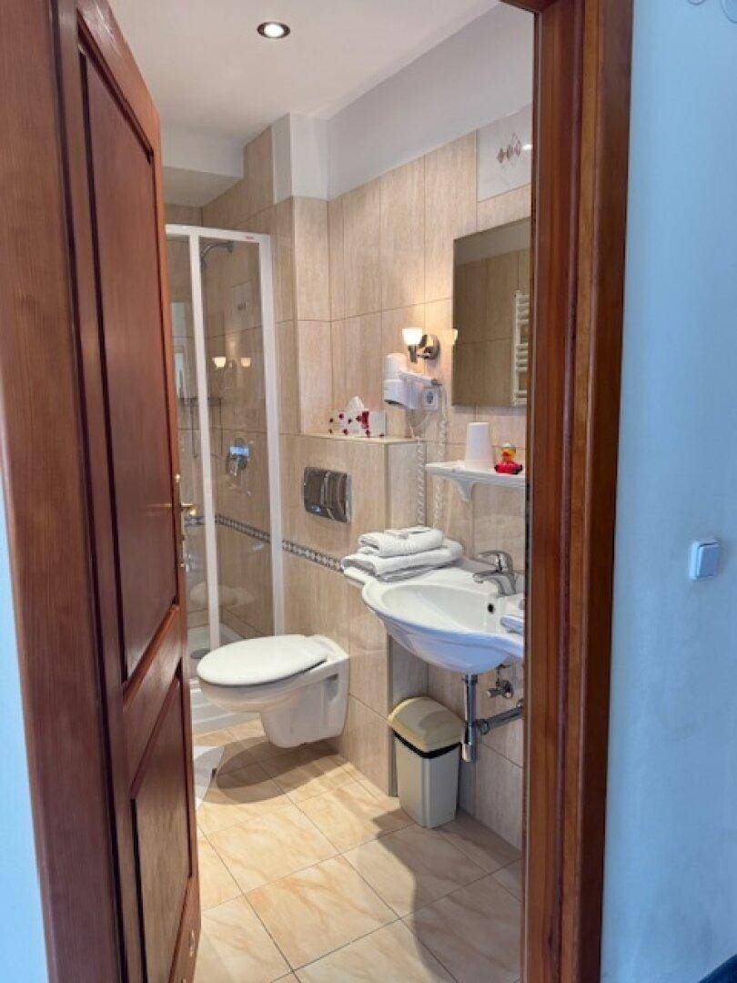 Kleines Badezimmer mit beigefarbenen Fliesen, Duschkabine, WC und Waschbecken, durch eine Holztür sichtbar.