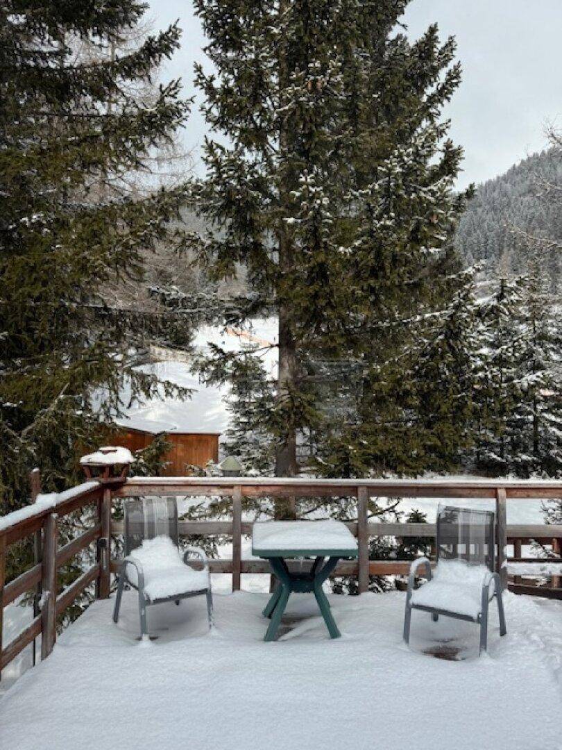 Kleiner Balkon mit Holzzaun und Gartenmöbeln, bedeckt mit Schnee und umgeben von verschneiten Bäumen.