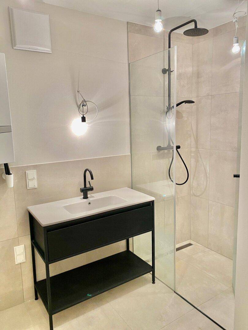 Modernes Badezimmer mit begehbarer Dusche, schwarzem Waschtisch und hellen Fliesen.