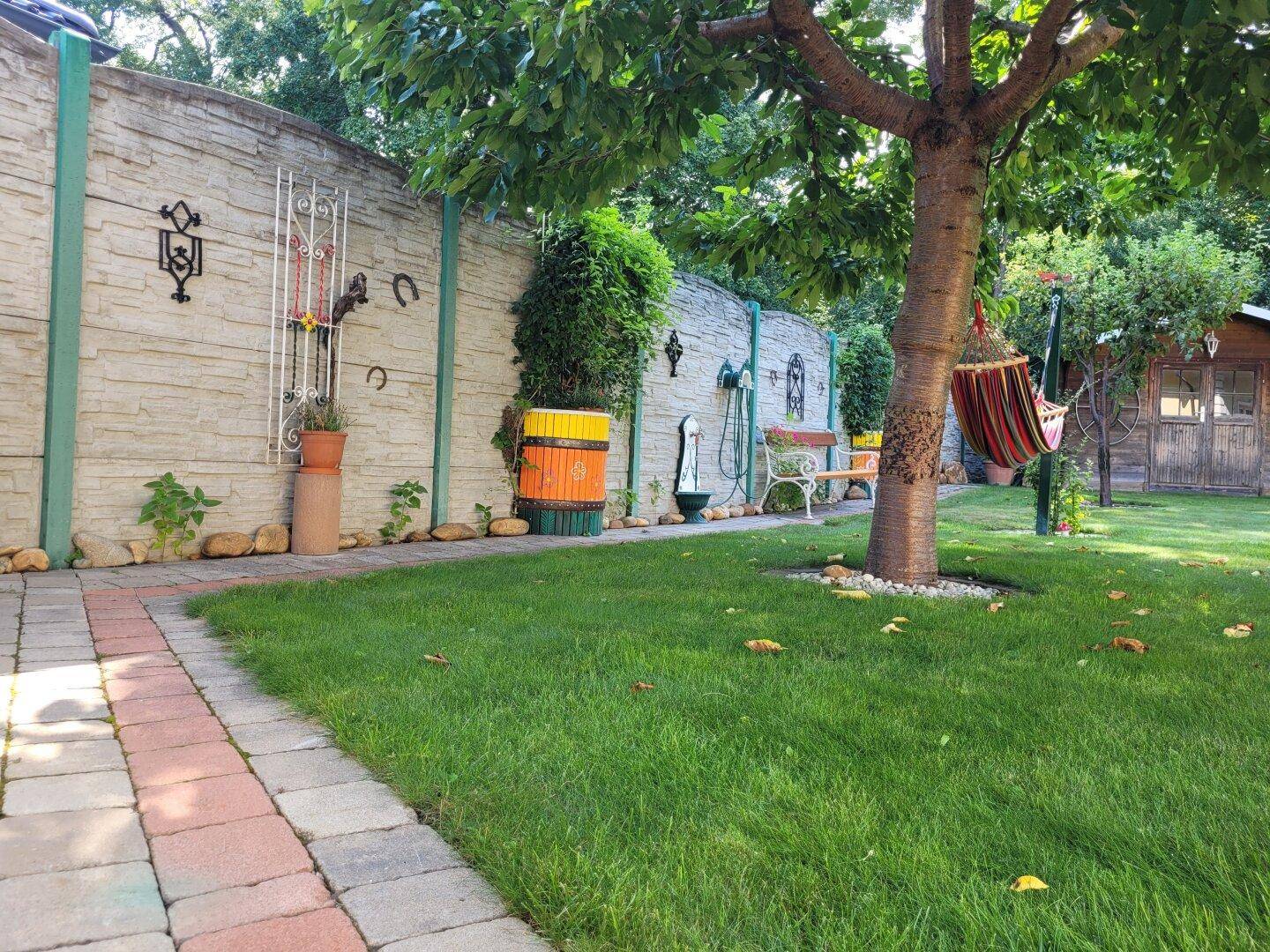 Gepflegter Garten mit Rasenfläche, Steinmauer und Hängematte unter Bäumen.