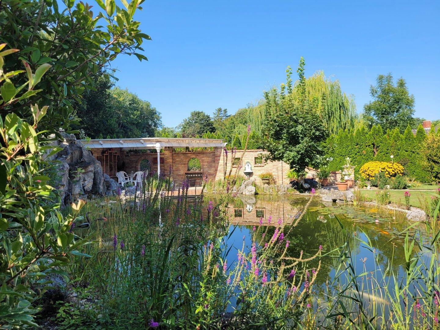 Großer Gartenteich mit üppiger Bepflanzung und einem Pavillon im Hintergrund.