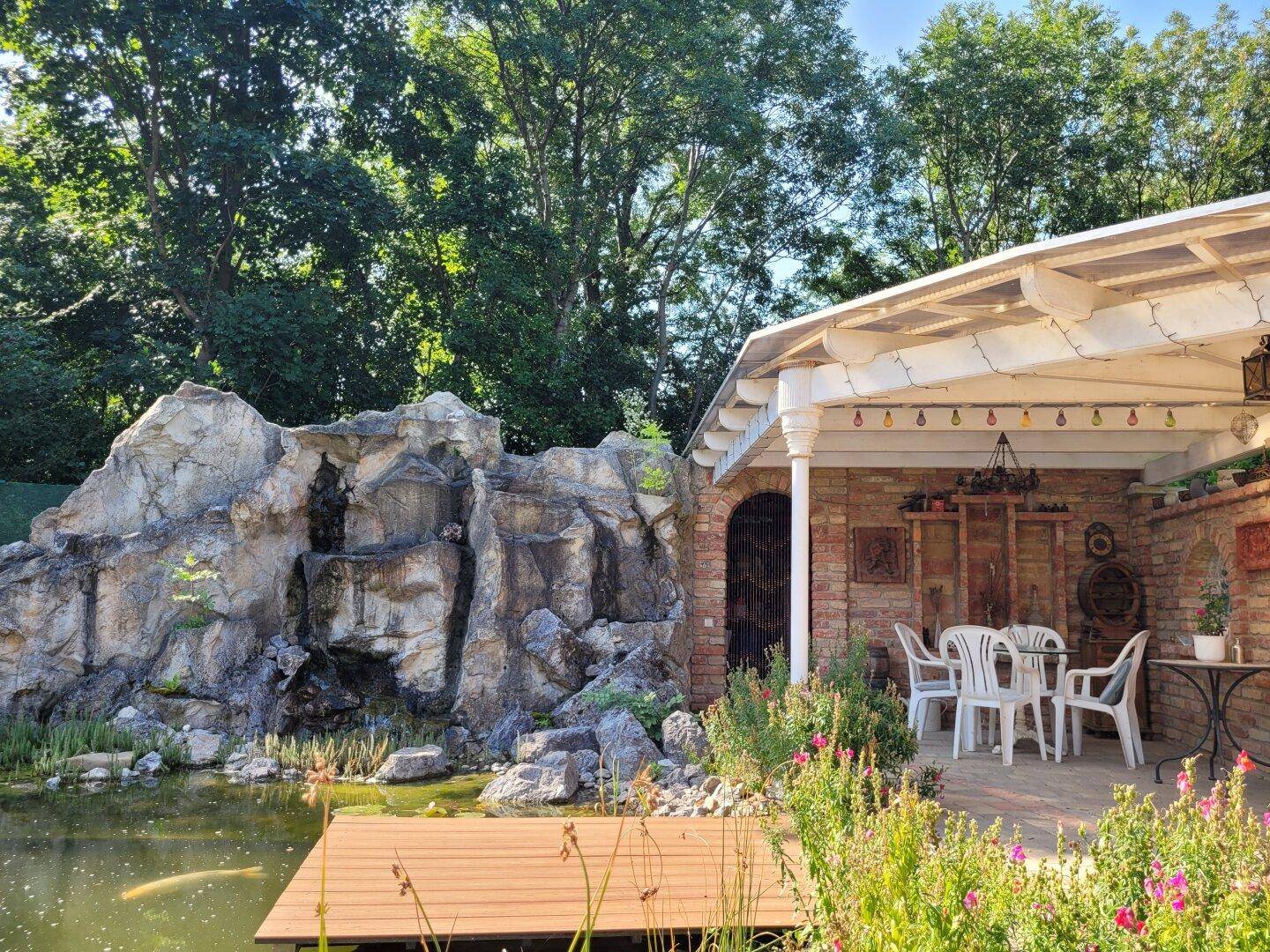 Gemütliche Terrasse am Teich mit Sitzgelegenheiten und einem dekorativen Felsen.
