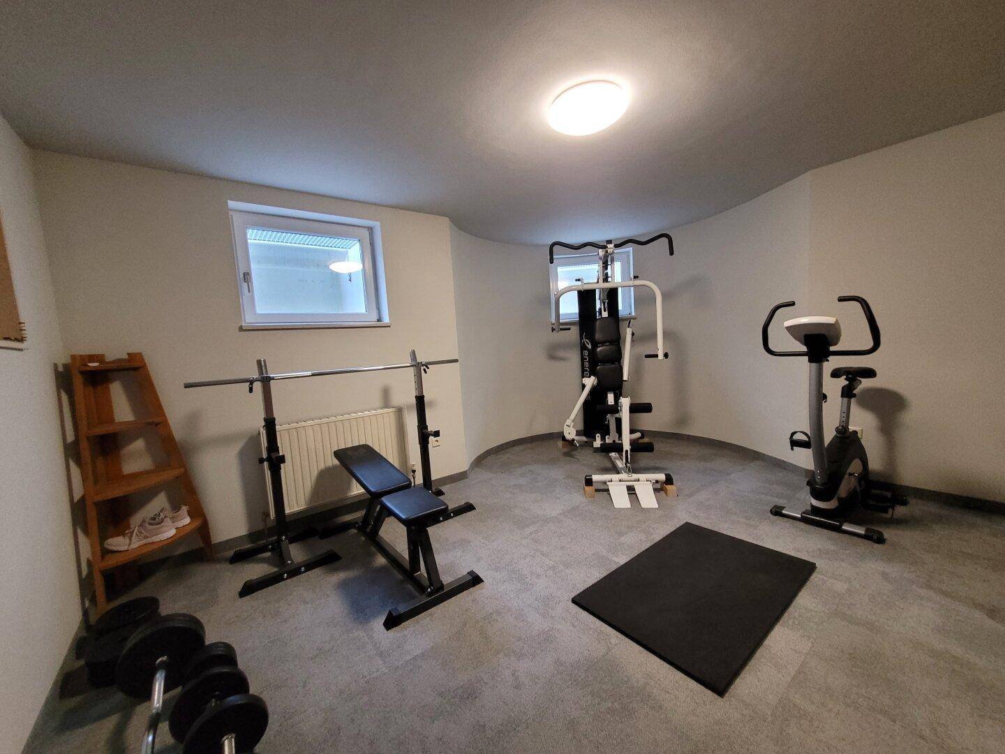 Geräumiger Fitnessraum im Untergeschoss mit verschiedenen Trainingsgeräten und einem Fenster für Tageslicht.