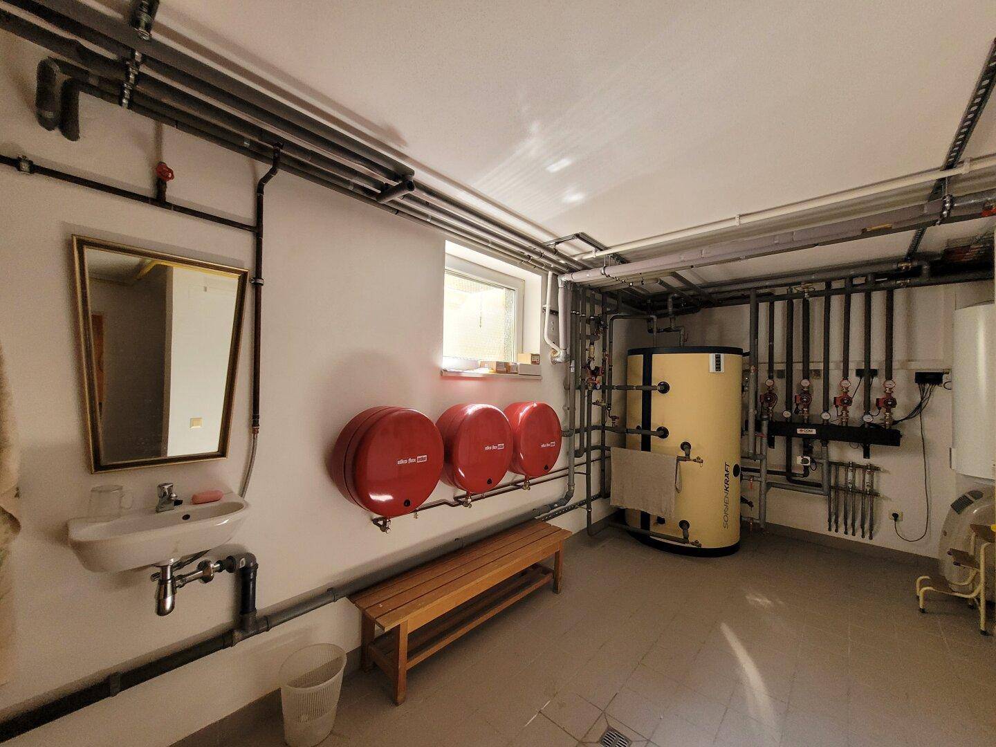 Technikraum im Keller mit Heizungsanlage, Warmwasserspeicher, freiliegenden Rohren und kleinem Waschbecken.