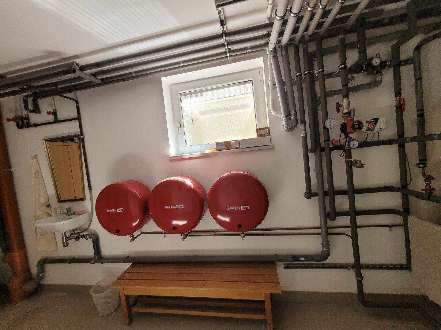 Detailansicht des Technikraums mit Waschbecken, Spiegel, Holzbank und Heizungsrohren an der Wand.