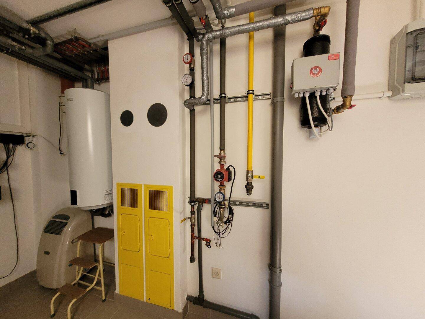 Weiterer Bereich des Technikraums mit Heizungsinstallationen, Warmwasserspeicher und gelben Schränken.