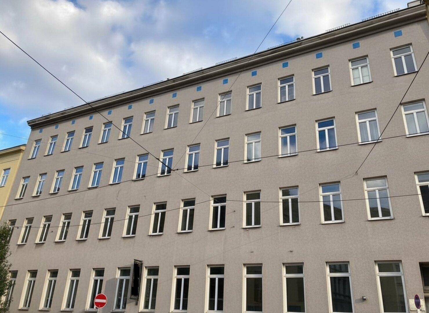 Graue Gebäudefassade mit vielen Fenstern und blauen Akzenten unter einem klaren blauen Himmel.